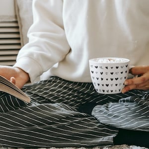 May include: A person wearing striped trousers and a white jumper sits on a white rug, holding a white mug with black hearts and a book. The mug contains a hot drink with marshmallows.