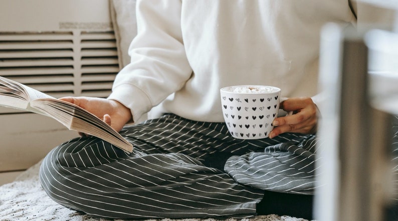 May include: A person wearing striped trousers and a white jumper sits on a white rug, holding a white mug with black hearts and a book. The mug contains a hot drink with marshmallows.