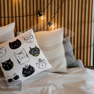 May include: A white pillow with a pattern of black and white cat faces, resting on a bed with other pillows. The cats have various expressions and accessories. A string of lights hangs above the bed, adding a warm glow.