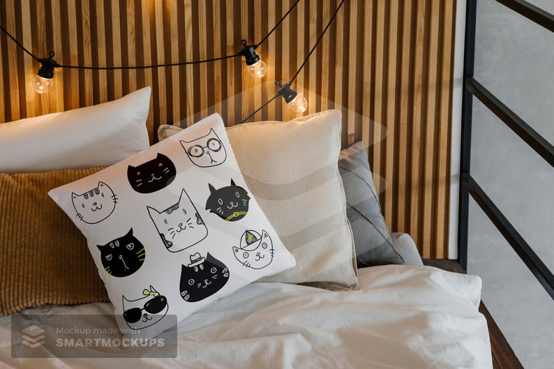 May include: A white pillow with a pattern of black and white cat faces, resting on a bed with other pillows. The cats have various expressions and accessories. A string of lights hangs above the bed, adding a warm glow.