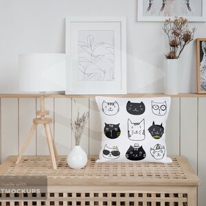 May include: White square throw pillow with a pattern of illustrated cat faces in black and white. The pillow is displayed on a wooden table with a white lamp and framed artwork in the background. The room has a neutral color palette.