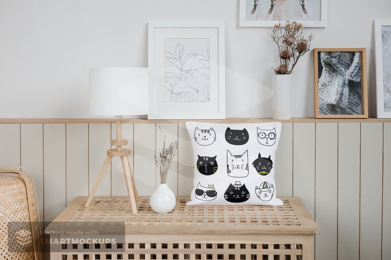 May include: White square throw pillow with a pattern of illustrated cat faces in black and white. The pillow is displayed on a wooden table with a white lamp and framed artwork in the background. The room has a neutral color palette.