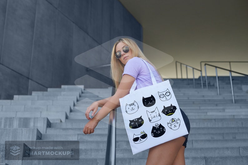 May include: White tote bag featuring a collection of illustrated cat faces in black and white. The bag is held by a person wearing a light purple t-shirt and sunglasses. The background includes concrete steps and a modern building.