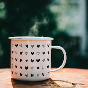May include: A white enamel mug with a black heart pattern, filled with a steaming beverage. The mug has a blue rim and a handle.