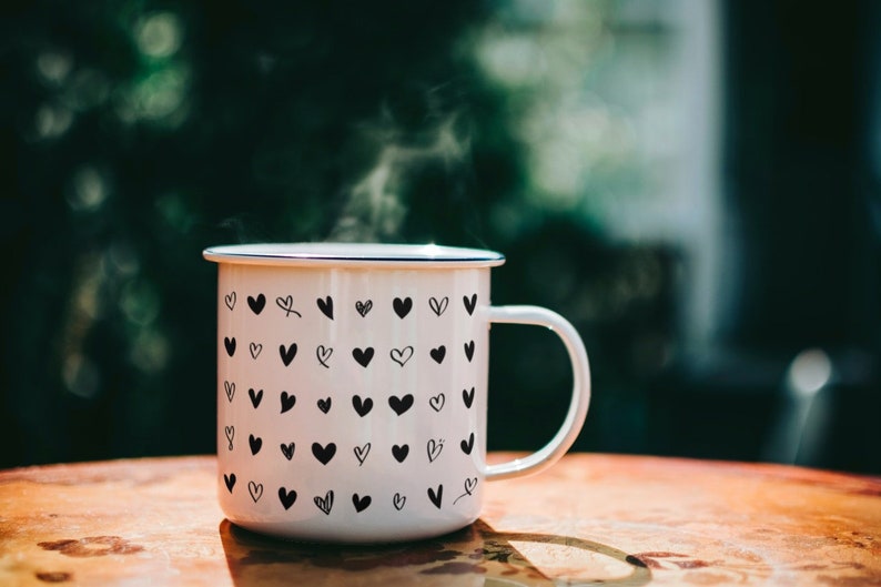 May include: A white enamel mug with a black heart pattern, filled with a steaming beverage. The mug has a blue rim and a handle.
