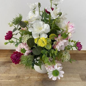 May include: A white ceramic vase filled with a colourful arrangement of artificial flowers. The bouquet includes white, pink, and burgundy flowers, as well as green foliage.