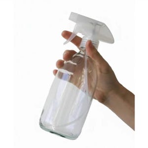 Könnte beinhalten: Eine transparente Glassprühflasche mit weißem Auslöser und Düse wird in einer Hand vor weißem Hintergrund gehalten. Die Flasche hat eine dreieckige Form und ist teilweise mit einer weißen Flüssigkeit gefüllt.