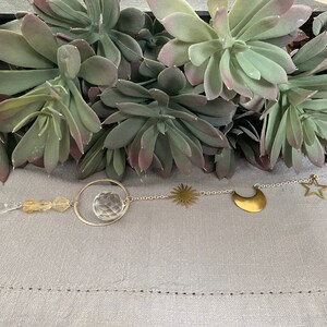 May include: A crystal suncatcher with a clear crystal orb, gold beads, and gold charms in the shape of a star, crescent moon, and sun. The suncatcher is hanging from a silver chain.