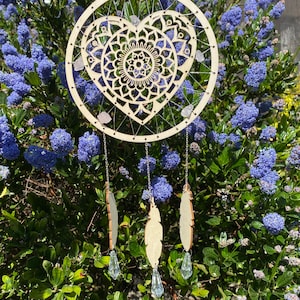 May include: A wooden dreamcatcher with a heart-shaped mandala design and crystals hanging from it. Three wooden feathers with crystal accents hang from the bottom of the dreamcatcher.