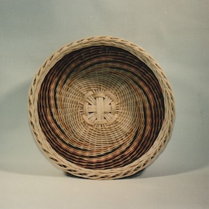 May include: A round woven basket with a brown and white pattern. The basket is made of natural materials and has a woven design.