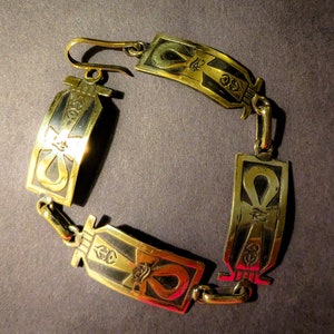 May include: A gold-toned bracelet featuring an ankh symbol design. The bracelet has a hook clasp and is made of metal.