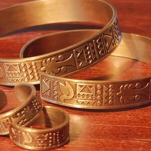 Può includere: Tre braccialetti in metallo dorato con intricati disegni geometrici. I braccialetti sono impilati uno sopra l'altro, con il braccialetto più grande in basso e il più piccolo in alto.