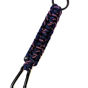 May include: A navy blue and red paracord keychain with a black carabiner clip and key ring. The braided cord has a textured appearance. The keychain is designed for attaching keys or other small items.