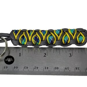 A gray and yellow braided keychain is being measured against a ruler to determine its length.