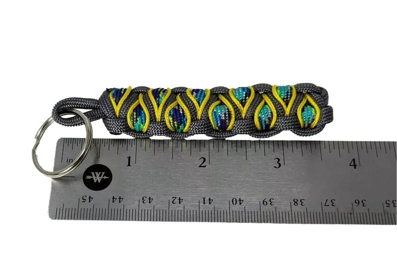 A gray and yellow braided keychain is being measured against a ruler to determine its length.