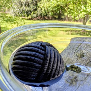 May include: A black decorative knot, likely a nautical item, rests inside a clear glass container. The knot is made of tightly woven rope, and the glass reflects the outdoor surroundings. The knot is a dark, solid color.