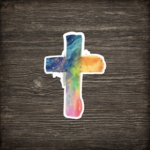 May include: A colorful sticker in the shape of a cross. The cross is filled with a vibrant mix of colors, including blue, orange, yellow, and pink, with a white border. The sticker is placed on a dark wood grain background.