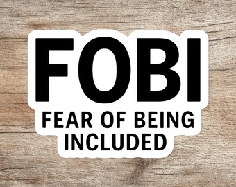May include: A white sticker with black text that reads "FOBI" above "FEAR OF BEING INCLUDED". The sticker is rectangular with rounded corners, set against a wood grain background.