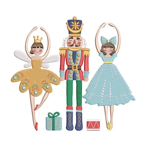 Könnte beinhalten: Gesticktes Design mit einem Nussknacker-Soldaten, flankiert von zwei Ballerinas. Der Nussknacker trägt eine rot-blaue Uniform mit goldener Krone. Eine Ballerina trägt ein gelbes Kleid und Flügel, die andere ein blaues Kleid. Ein Geschenk und eine Trommel sind ebenfalls enthalten.