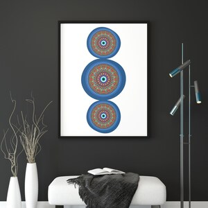 May include: A framed print featuring three blue and orange mandala designs with an evil eye motif. The print is hanging on a black wall above a white ottoman with a black throw blanket.
