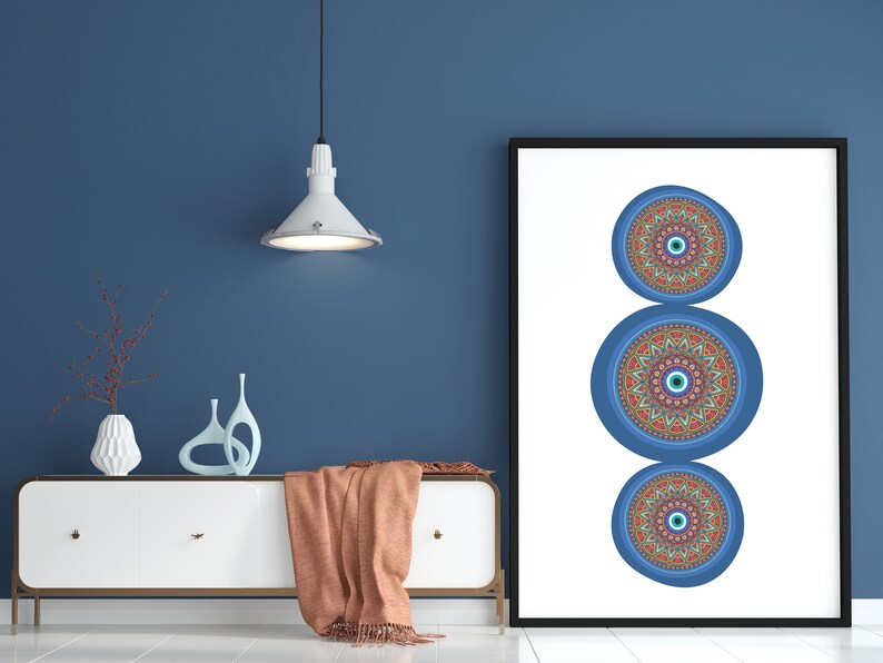 May include: A framed print featuring three blue and red mandala designs with an evil eye in the centre of each. The print is hanging on a blue wall above a white dresser with a brown throw blanket.