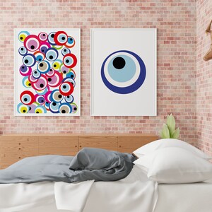 May include: Two framed prints of colorful evil eye designs. The print on the left features a cluster of overlapping evil eyes in various colors, while the print on the right features a single large blue evil eye with white and black accents. The prints are hanging on a brick wall above a wooden headboard and a bed with white and gray bedding.