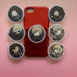 May include: A red phone case with seven character-themed acrylic charms. The charms feature cartoon illustrations of characters in a circular design. The background is a pink glittery surface.