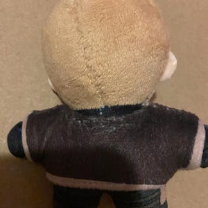 May include: A plush doll with a light brown head and a dark brown jacket. The doll has black trousers and a silver chain attached to the top of its head. The jacket has a white trim.