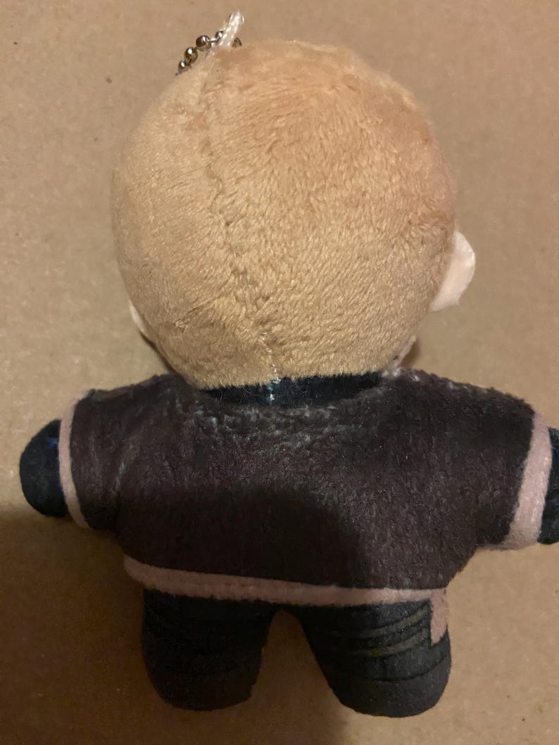 May include: A plush doll with a light brown head and a dark brown jacket. The doll has black trousers and a silver chain attached to the top of its head. The jacket has a white trim.