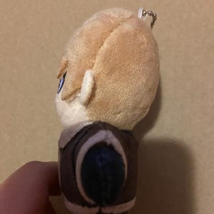 May include: A plush doll with light brown hair and blue eyes. The doll is wearing a brown jacket with a white collar. A silver keychain is attached to the top of the doll's head.