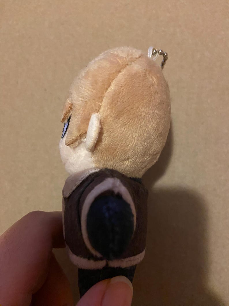 May include: A plush doll with light brown hair and blue eyes. The doll is wearing a brown jacket with a white collar. A silver keychain is attached to the top of the doll's head.