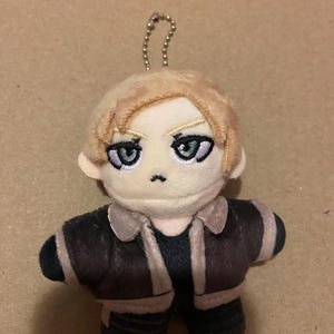May include: A plush doll with light brown hair and blue eyes, wearing a brown jacket with tan trim and black trousers. The doll has a small silver chain attached to the top of its head.