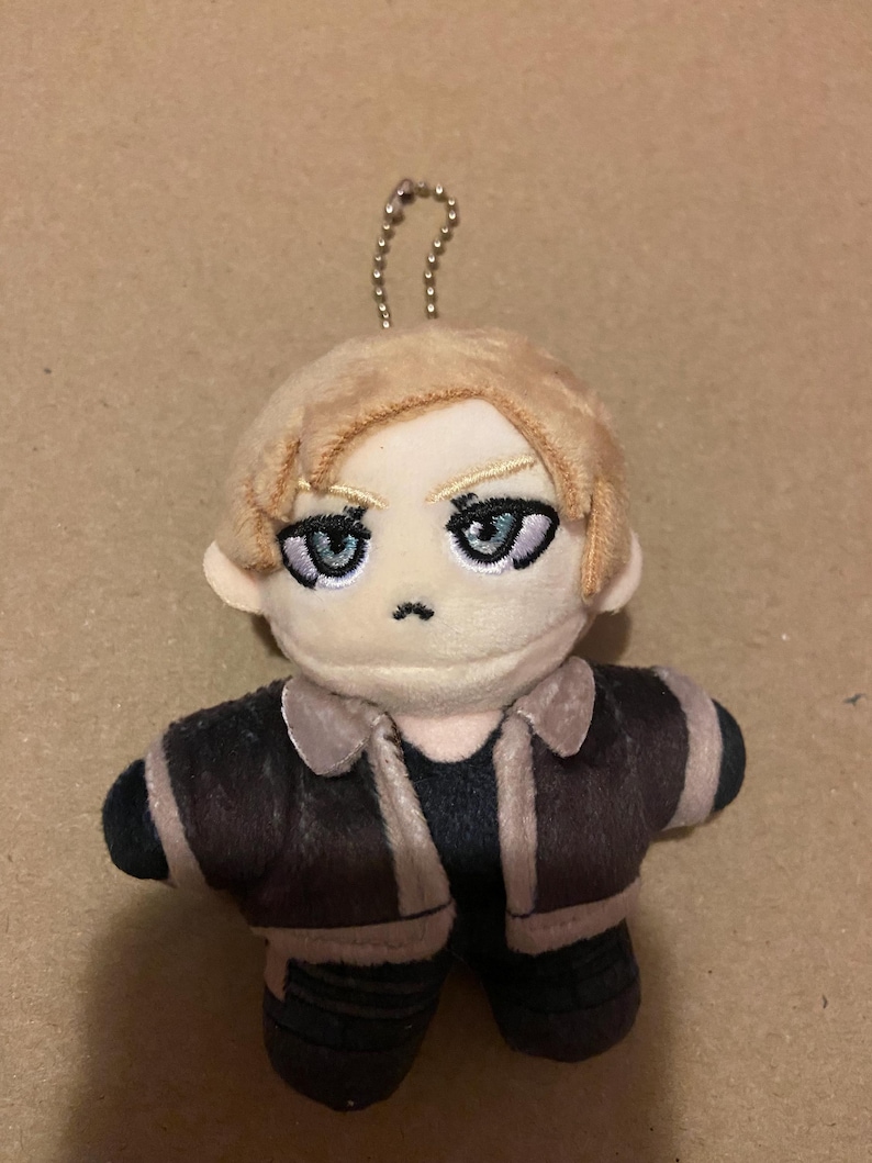 May include: A plush doll with light brown hair and blue eyes, wearing a brown jacket with tan trim and black trousers. The doll has a small silver chain attached to the top of its head.