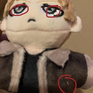 May include: A plush doll with a tan face, blonde hair, and a brown jacket with tan trim. The word "DEFECTIVE" is written in red at the top of the image. The doll has blue eyes and a black shirt.
