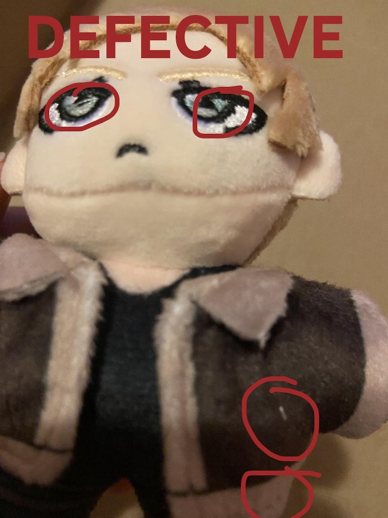 May include: A plush doll with a tan face, blonde hair, and a brown jacket with tan trim. The word "DEFECTIVE" is written in red at the top of the image. The doll has blue eyes and a black shirt.