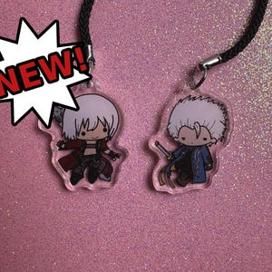 May include: Two clear acrylic charms featuring cartoon characters with white hair and detailed outfits. One charm has a red coat, and the other has a blue coat. Both charms are attached to black cords. A white starburst with the word "NEW!" is in the image.