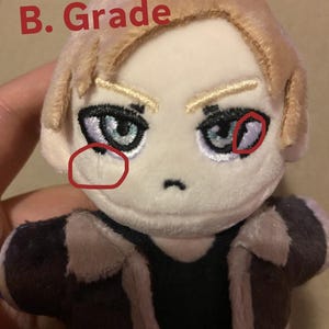 May include: A plush doll with blonde hair and detailed facial features, including blue eyes and eyebrows. The doll is wearing a dark jacket with a light brown collar. The text "B. Grade" is written in red on the top left.
