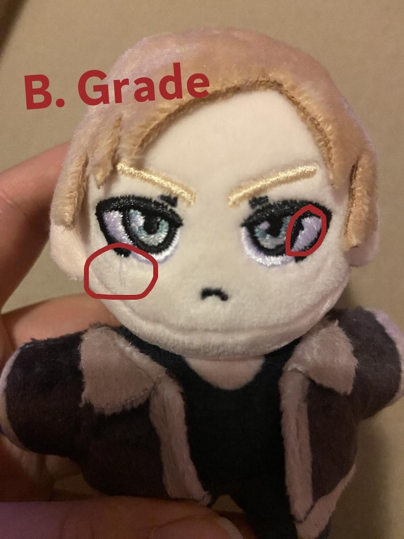 May include: A plush doll with blonde hair and detailed facial features, including blue eyes and eyebrows. The doll is wearing a dark jacket with a light brown collar. The text "B. Grade" is written in red on the top left.