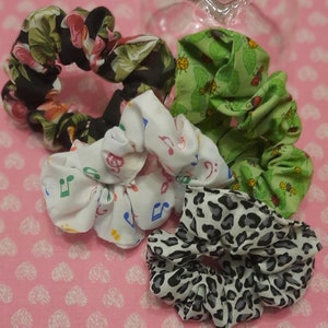 May include: Four colourful scrunchies made of fabric. The scrunchies are in a variety of patterns, including floral, musical notes, and a green leafy pattern with ladybirds. The scrunchies are arranged on a pink background with a white heart pattern.