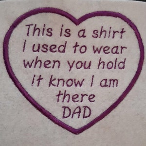 May include: A heart-shaped fabric patch with a purple embroidered border and purple embroidered text that reads: This is a shirt I used to wear when you hold it know I am there DAD. The patch is made of a light cream-coloured felt material.