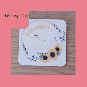K&ouml;nnte beinhalten: Ein quadratischer Untersetzer in Wei&szlig; mit einem Kranz aus blauen Eukalyptusbl&auml;ttern und gelben Sonnenblumen. Eine Biene mit schwarzen und gelben Streifen fliegt im Kreis um den Text "BEE HAPPY".