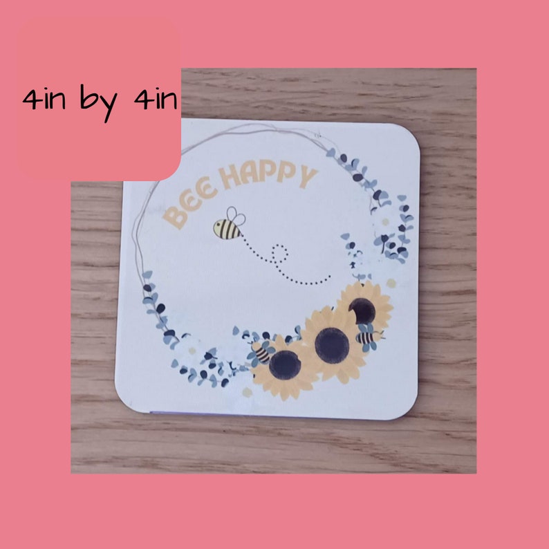 K&ouml;nnte beinhalten: Ein quadratischer Untersetzer in Wei&szlig; mit einem Kranz aus blauen Eukalyptusbl&auml;ttern und gelben Sonnenblumen. Eine Biene mit schwarzen und gelben Streifen fliegt im Kreis um den Text "BEE HAPPY".