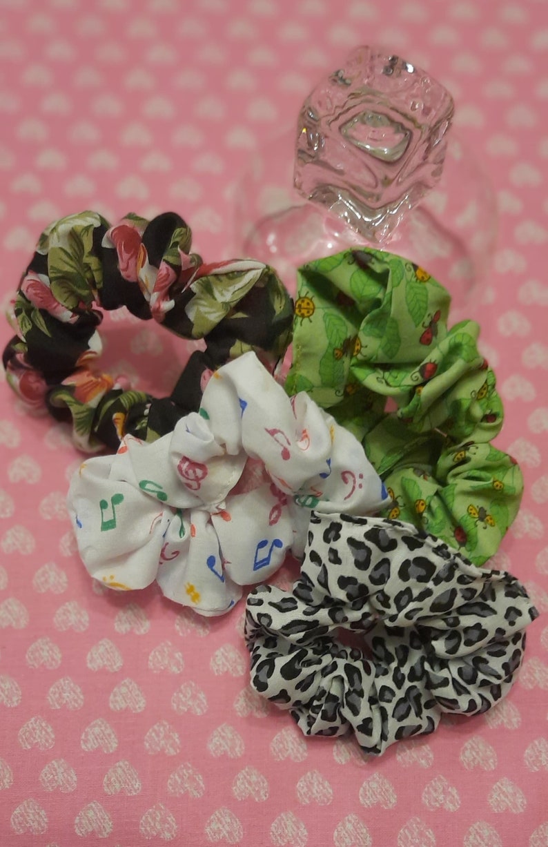 May include: Four colourful scrunchies made of fabric. The scrunchies are in a variety of patterns, including floral, musical notes, and a green leafy pattern with ladybirds. The scrunchies are arranged on a pink background with a white heart pattern.