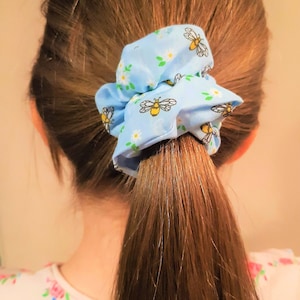 May include: A light blue scrunchie with a white floral and bee pattern. The scrunchie is tied around a ponytail.
