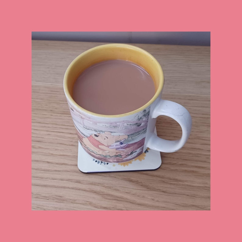 K&ouml;nnte beinhalten: Eine gelbe und wei&szlig;e Keramiktasse mit einem Winnie-the-Pooh-Motiv, gef&uuml;llt mit einem braunen Getr&auml;nk, auf einer Holzoberfl&auml;che.