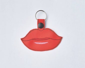 May include: A red lip-shaped keyring featuring a smooth texture with dark stitching along the edges. The design includes a black snap button at the top, a silver metal split ring for keys, and embroidered details representing the mouth and teeth.