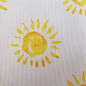 May include: White fabric with a repeating pattern of yellow suns. Each sun has a yellow centre and orange rays.