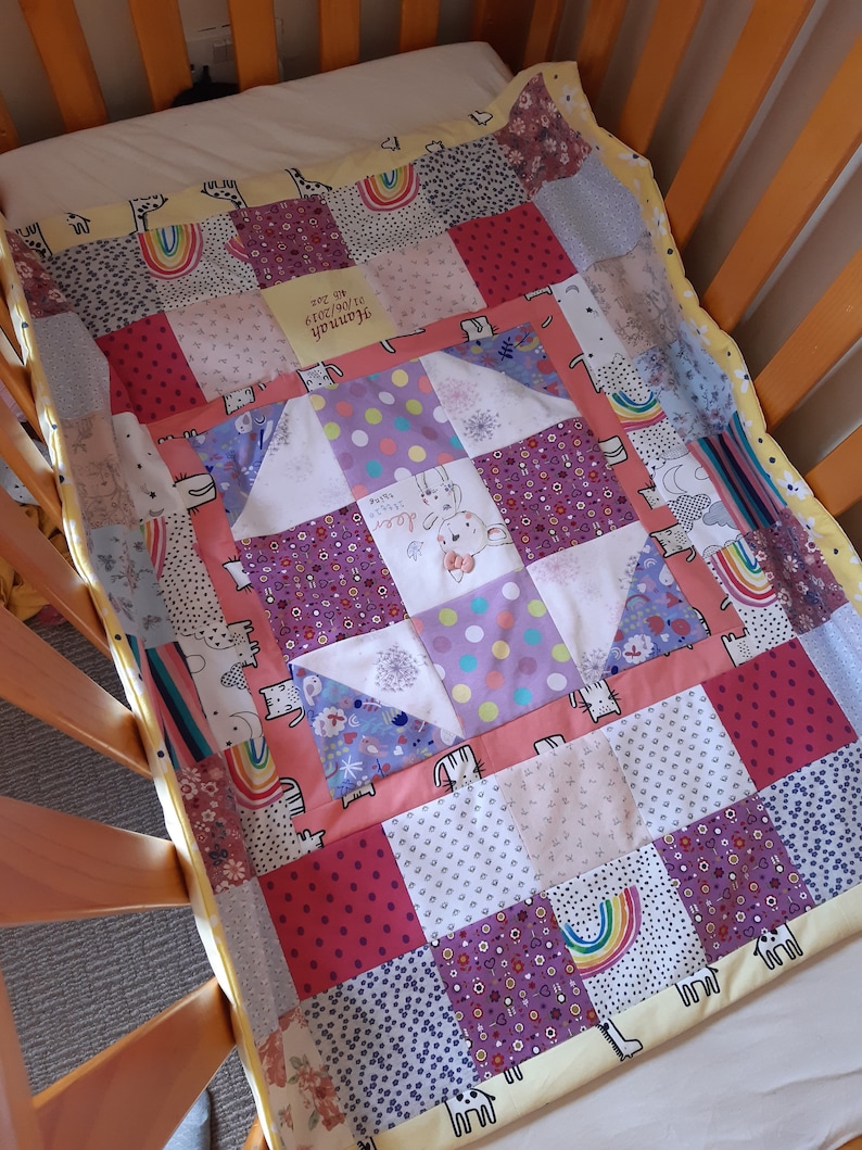 Puede incluir: Una colcha de beb&eacute; de retazos en colores rosa, morado y amarillo. La colcha presenta una variedad de patrones, incluyendo lunares, rayas e estampados florales. La colcha est&aacute; hecha de tela suave y c&oacute;moda, perfecta para acurrucarse con un peque&ntilde;o. La colcha tiene un mensaje personalizado que dice "Hannah, te queremos mucho, 01.08.2019".