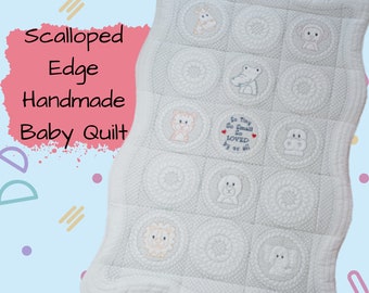 May include: A white baby quilt with a scalloped edge. The quilt is made of soft fabric and features a variety of embroidered animal designs, including a lion, giraffe, monkey, crocodile, hippopotamus, and dog. The quilt also includes the text "So Tiny So Small So Loved By Us All".