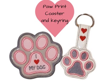 May include: A paw print coaster and keyring set. The coaster is grey with pink paw pads and the text "I ❤️ MY DOG". The keyring is white with red heart accents and a paw print design.